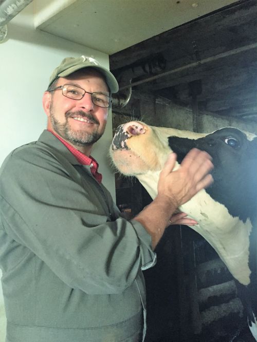 A smiling man stands in front of a dairy cow while scratching under the cow's chin.